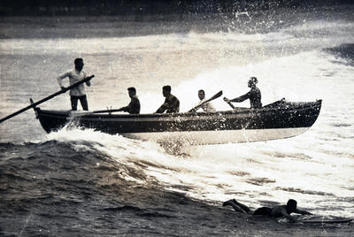 Surf-boat crashing through a wave and a man on a surf-board nearby