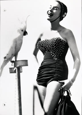 Female wearing 'sports de jour fashion' bathing costume with beaded bodice, beaded bathing cap with chinstrap, white cockatoo on stand on left.