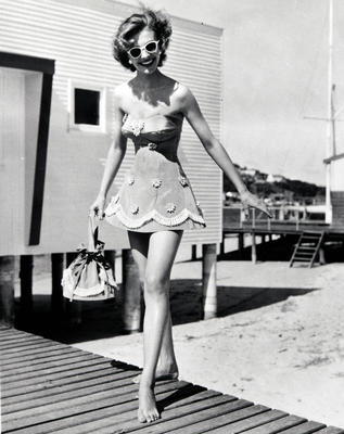 Female wearing 'sports de jour fashion' strapless bathing costume with white flower trim and tassle trim on bottom holding matching bag.