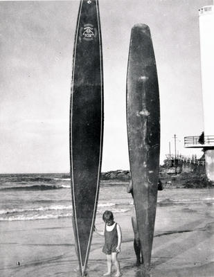 Child in front of two surfboards held up by members of Manly Life Saving Club Shark Tower on R.H.S also walkway to Fairy Bower.