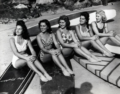 5 girls in swimwear sitting on long wooden boards on beach.