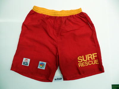 Long nylon board shorts in red with yellow waistband and printed "SURF RESCUE' on left leg - There is a hip pocket on each side + 'Kellogg's NUTRIGRAIN' + 'SURF LIFESAVING AUSTRALIA' on right leg. Unlined