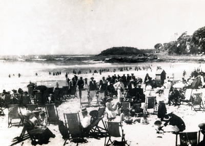 South-east across the southern end of Manly Beach towards Shelly Beach showing a number of people standing or sitting in deck-chairs with others in the surf or on the Fairy Bower walkway